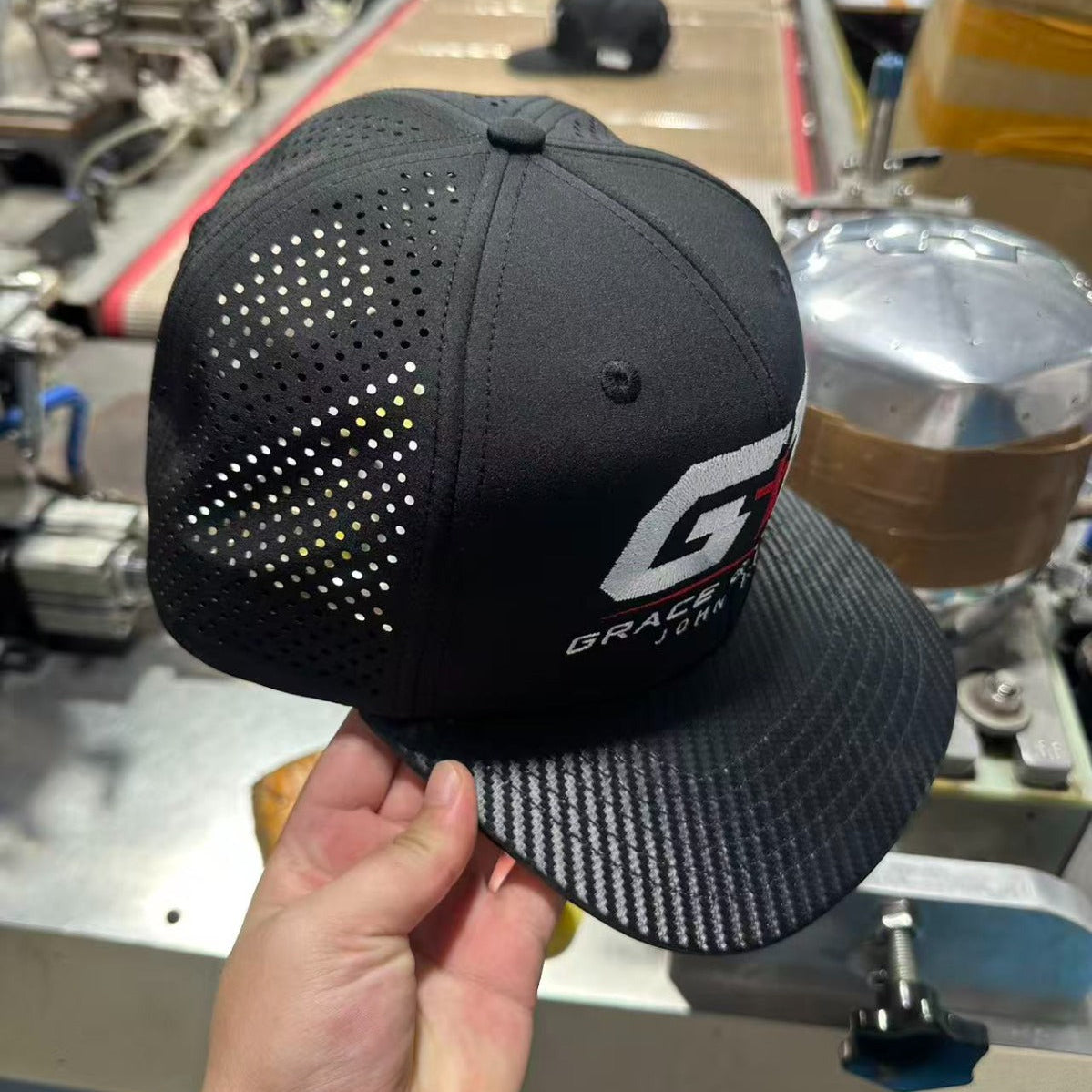A black trucker-style baseball cap with a perforated back panel and a carbon-fiber-look brim, held by a hand in a workshop setting. The front of the cap features a white and red 'G' logo with 'GRAP.