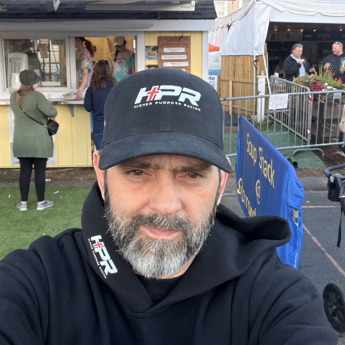 A man with a gray beard wearing a black HPR cap and black hoodie takes a selfie in front of the Storrowton Soup Shack food stand at an outdoor market.