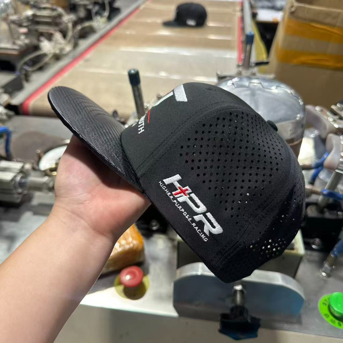 A black perforated baseball cap with white and red 'HSP' branding, held up by a hand in a busy workshop or factory setting.