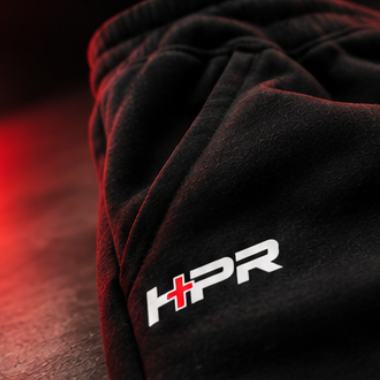 Close-up of black sweatpants with a white and red HPR logo on the thigh, photographed in dramatic red lighting against a dark background.
