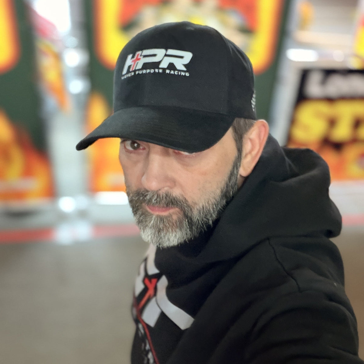 A man wearing a black baseball cap with a white and red 'HPR High Performance Racing' logo, paired with a black hoodie, photographed in a brightly lit indoor setting with blurred orange and yellow signage in the background.