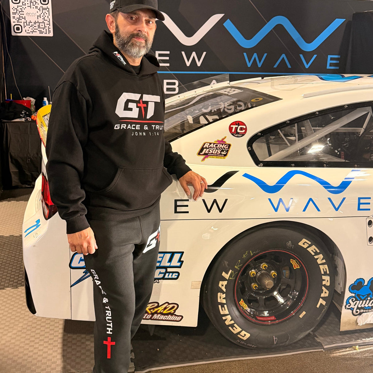 Man wearing the black Team Grace & Truth sweatsuit beside a race car